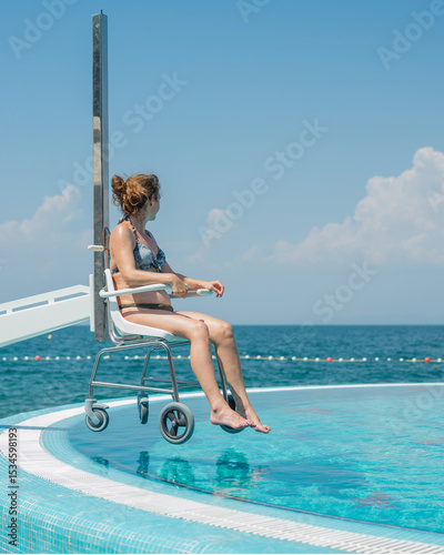 Lifeguard operating a pool chair lift to lift the smiling woman with disability to and from the pool safely during her summer vacation and sea hotel stay.