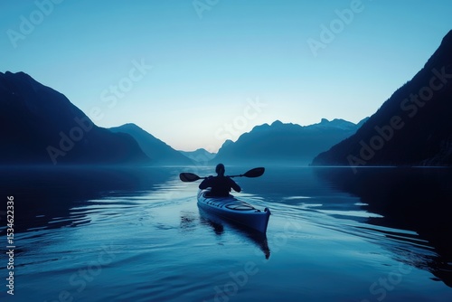 A lone kayaker paddles through calm waters at dusk, surrounded by majestic mountains under a serene, blue sky.