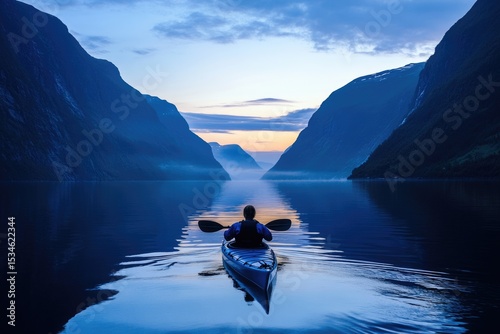 A kayaker paddles on a calm, blue lake surrounded by large, majestic mountains at twilight or early morning.