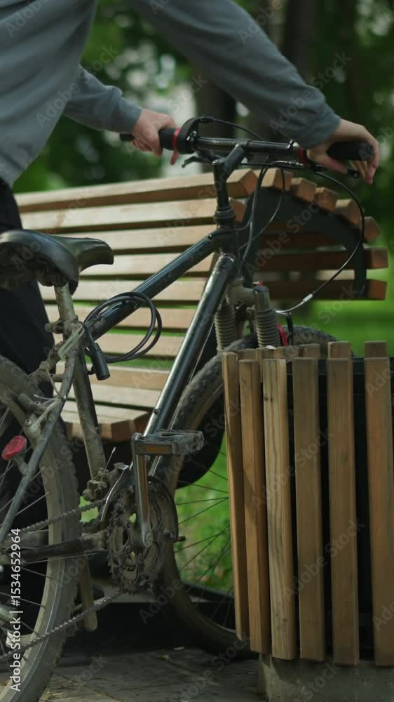 Young boy rides his bicycle into a peaceful park and parks it between a wooden bench and a nearby structure, he lets the bicycle rest on the bench as he sits down, surrounded by greenery and trees