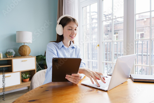 Young Woman Engaged in Remote Learning While Using a Tablet and Laptop at a Cozy Workspace in a Bright, Modern Apartment