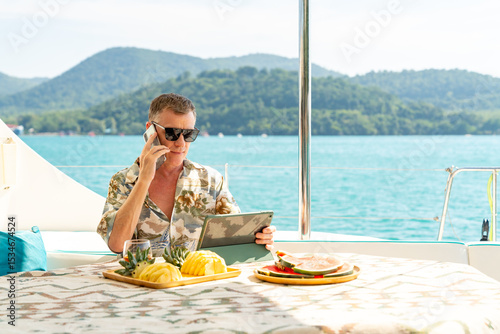 Caucasian businessman working on digital tablet and speaking on mobile phone for online corporate business on luxury catamaran boat yacht sailing in ocean. Workation on summer beach holiday vacation.