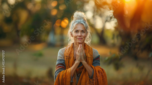Smiling mature woman in prayer pose with orange shawl in serene autumn park at sunset