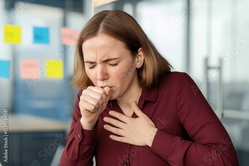 Coughing: A woman in a maroon blouse is coughing while sitting at an office desk, holding her chest in discomfort