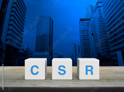 CSR acronym on white block cubes on wooden table over modern office city tower and skyscraper, Corporate Social Responsibility concept