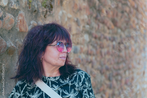 Stylish senior woman wearing sunglasses and looking away while posing in front of an old stone wall