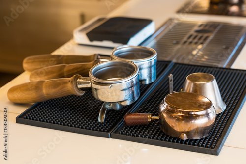 Coffee shop counter with wooden-handled portafilters, copper milk jug, and tamper showcased on a rubber mat near a sink
