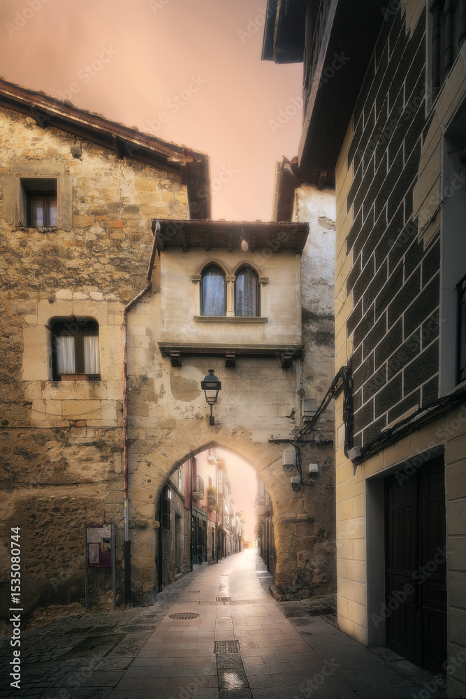 Fototapeta premium Historic stone archway and narrow street in Medina de Pomar, Burgos, Spain, at sunrise. Medieval architecture and charming old town atmosphere