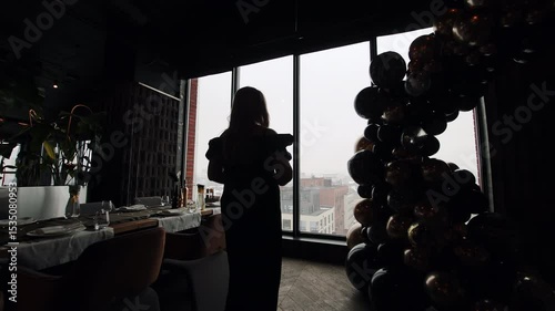Woman at elegant indoor event with city view. Silhouetted woman stands in upscale venue near a balloon arch and decorated table, overlooking a cityscape through.