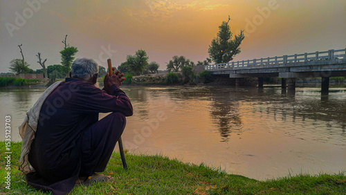 Elderly man contemplating quietly by the riverside during sunset in Pakistan, symbolizing wisdom, reflection, solitude, cultural identity, and peaceful connection with nature