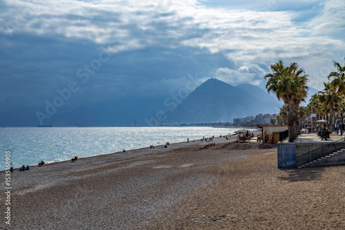 Fototapeta Naklejka Na Ścianę i Meble -  Konyaaltı's pebble beach stretches into distance under mixed March weather: dark clouds haze mountains while sun sparkles on sea. Palm trees and port cranes frame the scene. Antalya, Turkey.
