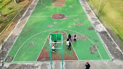 Aerial view of a worn green basketball court with white lines circles and people standing near a hoop