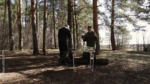 Two people enjoy a peaceful morning in the forest, preparing breakfast at a small folding table while surrounded by tall pine trees and dappled sunlight filtering through the canopy
