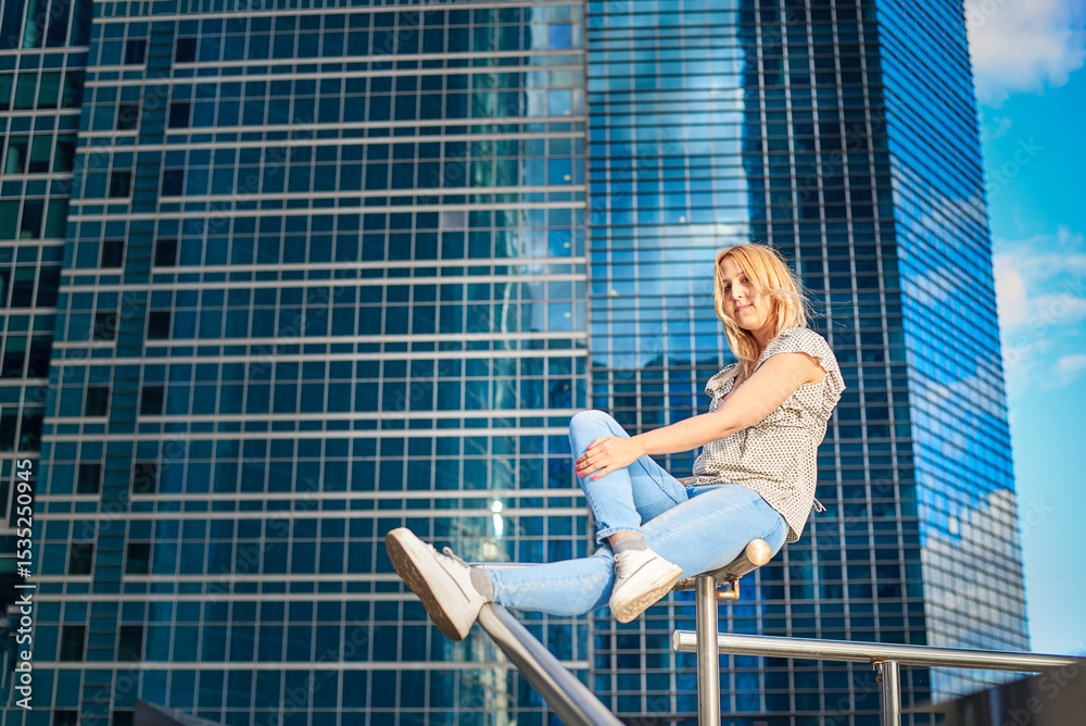 Fototapeta premium A woman with a beautiful figure leans on a railing in front of a tall glass building. She is wearing a white shirt and blue jeans.
