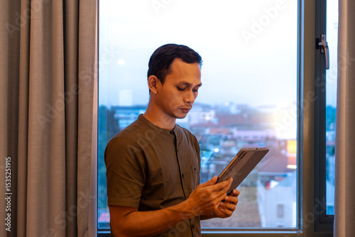 Asian Man using tablet near window with city view in the background Free Download Asian Man using tablet near window with city view in the background - GFXLoad