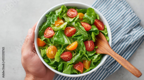 Fresh green salad with crisp lettuce, juicy red and yellow cherry tomatoes in white bowl, hand holding bowl, wooden spoon, striped cloth, healthy and vibrant meal