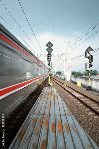A blur of a train passes a station platform with tracks and railway signals under a cloudy sky, hinting at the bustle of North Jakarta's transportation. photo