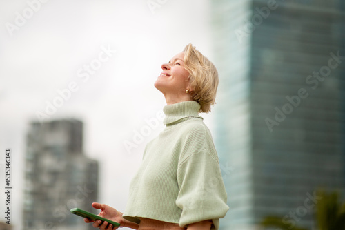 Canvastavla Mature woman with blond hair looking up and smiling while holding phone outdoors