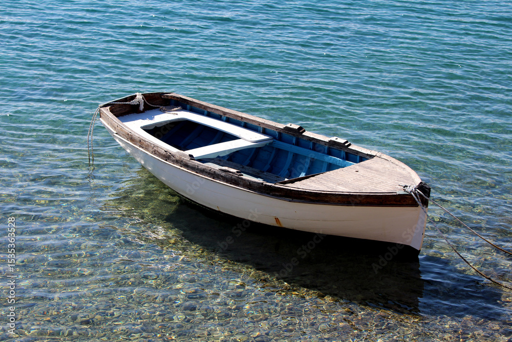 Naklejka premium White and blue wooden rowboat with dark trim floats in shallow, crystal-clear water, tethered by ropes at the bow and stern, revealing pebbles beneath and gentle ripples in bright sunlight