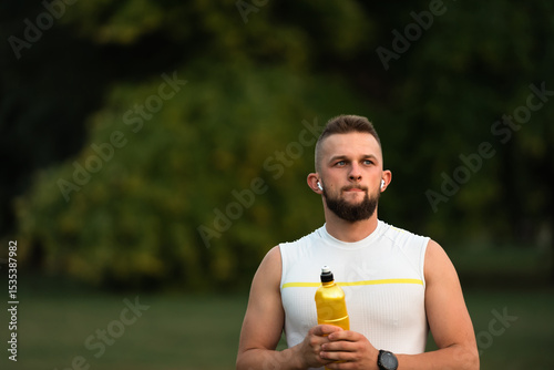 Photos A young, fit man listens to music on wireless earbuds and relaxes after a run or workout in a summer park, showcasing an active, healthy lifestyle