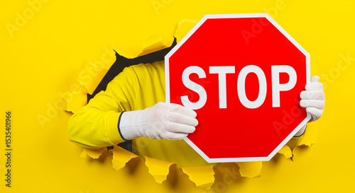 Person holding stop sign breaking through yellow paper background with gloved hands visible in frame