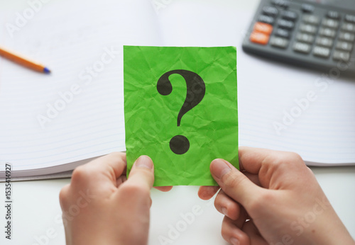 A student  holding a green paper note with a question mark on it in his hands over an open notebook and a calculator. Close up.