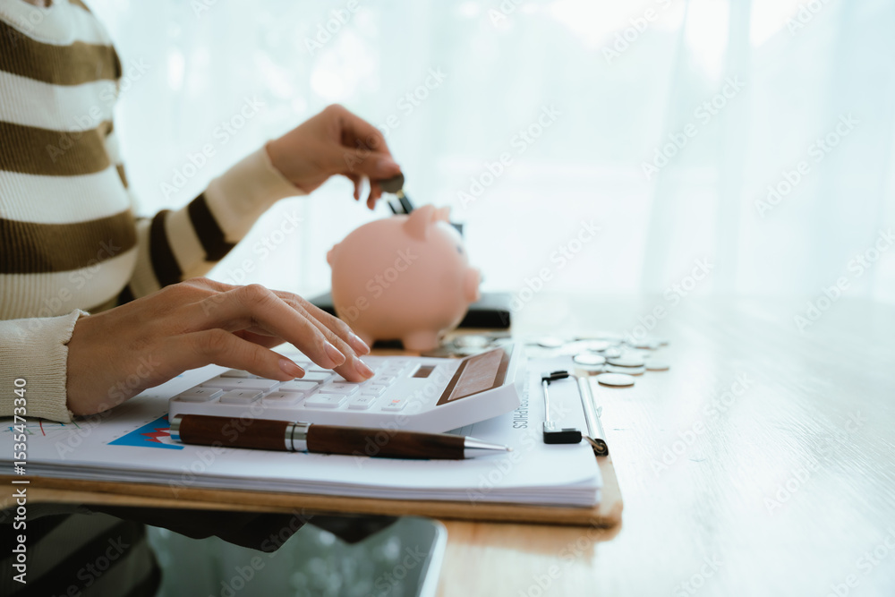 Fototapeta premium An Asian woman is putting money in a piggy bank while looking at a calculator, giving advice on financial planning.