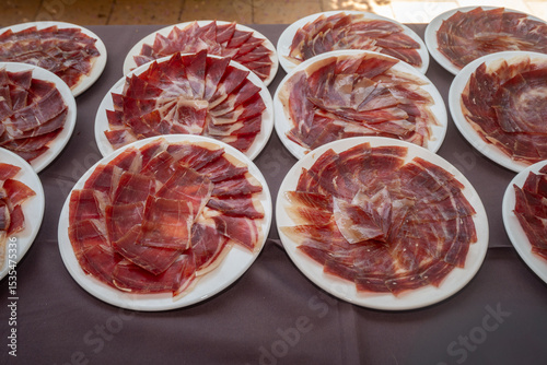 Mehrere weiße Teller, sorgfältig belegt mit hauchdünn geschnittenem spanischem Serrano-Schinken, sind auf einem gedeckten Tisch arrangiert. Die kunstvolle Anordnung der rotbraunen Schinkenscheiben