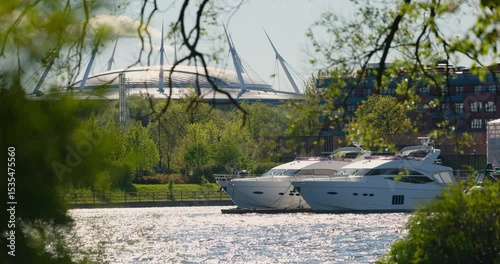 View of the mega yachts standing at the pier through the branches of trees on a clear sunny day, residential buildings and a stadium in the background, Russia, St.Petersburg
