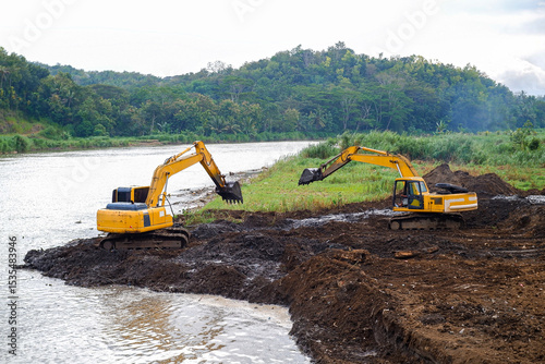 Excavators Operating Along Riverside in Natural Green Landscape. Excavators actively performing earth-moving work near a riverside amidst a green forested area.