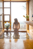 A woman enjoys a serene bath in a wooden tub by a panoramic window. Surrounded by flowers and soft daylight, the moment captures peaceful self-care and quiet luxury