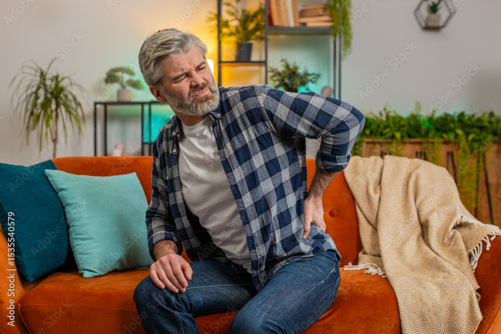 Fototapeta premium Tired senior bearded man suffering from back pain sitting on sofa in living room at home. Sad old male massages painful lower back muscles. Caucasian mature guy exhausted overworked in apartment.