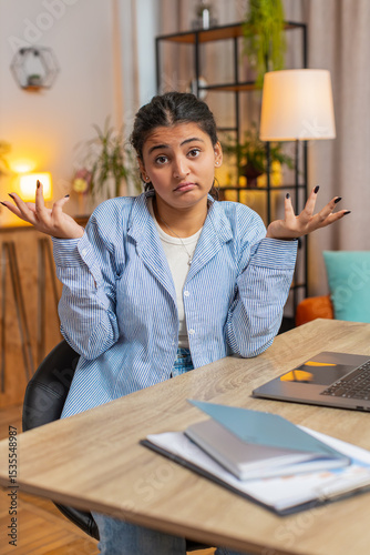 I dont know what to say. Confused woman feeling embarrassed about ambiguous question, having doubts, no answer idea, being clueless, uncertain. Indian girl sitting at home office desk table. Vertical.