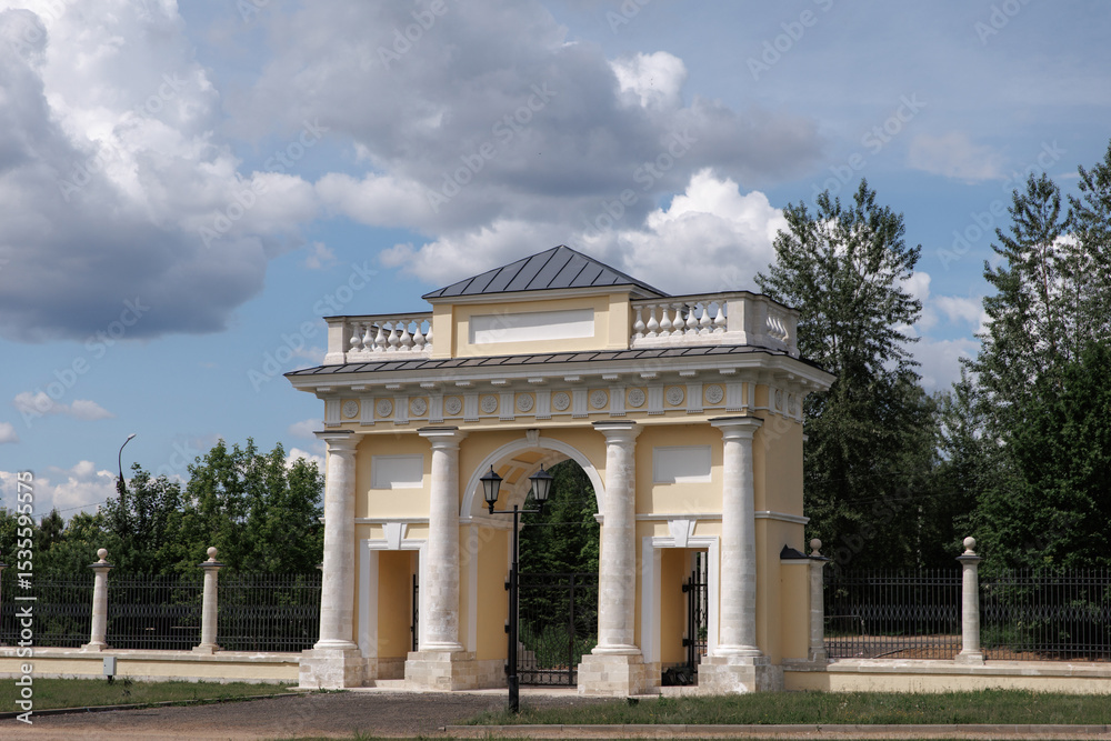 Obraz premium Captivating image of an elegant historic archway, framed by a vintage lamp post under a partly cloudy sky. Stunning architectural details and lush landscape provide a timeless charm.