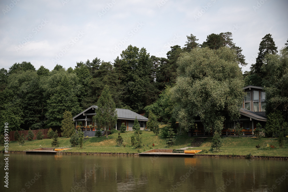 Obraz premium Peaceful lakeside scene with charming cottages nestled among lush greenery. The tranquil setting features a calm lake, dense forest, and inviting docks perfect for relaxation.