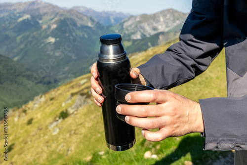 man pouring tea from thermos against mountain range background, hiking in mountains 