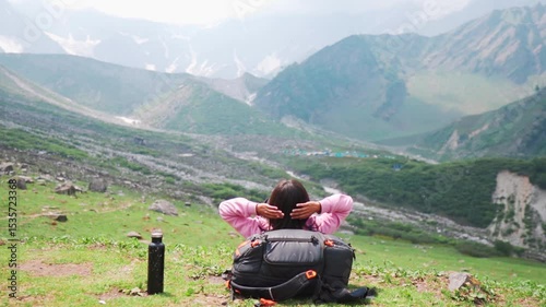 Relaxed teenager Indian girl lying on grass, resting head on bag pack and enjoying view of mountains during Beas kund hike in Himachal Pradesh, India. Female hiker relaxing in mountains. 
