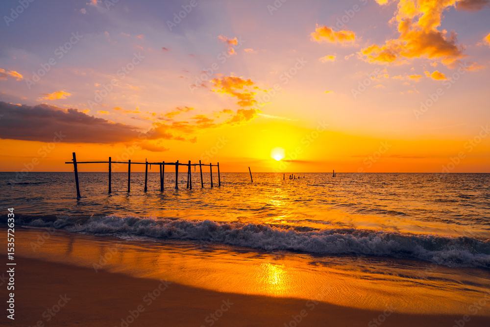 The view of the old wooden bridge decayed in the middle of the ocean in the evening. The sun is setting, making the sky a golden yellow. The waves hit the shore with orange light. There is copy space. The view of the old wooden bridge decayed in the middle of the ocean in the evening. The sun is setting, making the sky a golden yellow. The waves hit the shore with orange light. There is copy space.