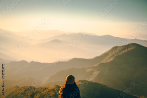 A woman standing on a mountain with sunrise in a soft tone. She turned around and looked at the beautiful view. There is a forest below with warm light.  Idea for nature wallpaper with copy space.