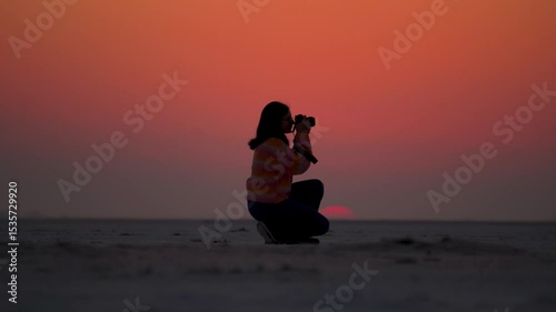 Silhouette of Indian girl holding camera and capture photos white sitting at White Desert of Kutch, Gujarat, India. Tourist exploring Kutch Desert during sunset. Big orange sun setting at horizon.