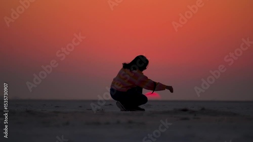 Sunset silhouette of teenager Indian girl grabbing salt crystals, desert sand from White Rann of Kutch in Gujarat, India. Travel, tourism and holidays concept. Tourist exploring nature. 