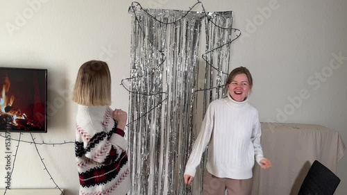 Two cheerful women in winter sweaters dance joyfully in front of a silver tinsel backdrop.  A cozy fireplace scene plays on a nearby TV, setting a festive mood. Perfect for holiday or celebration
