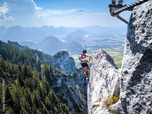 Climber On Fingersteig Via Ferrata Near Tegelberg: Adventurer Traversing Narrow Ridge With Harness And Helmet Against A Stunning Alpine Backdrop In Bavaria, Germany