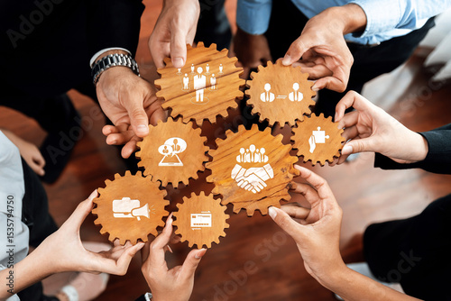 A group of diverse individuals gathers to hold wooden gears featuring business icons, symbolizing teamwork and collaboration in a dynamic workspace environment. Amity