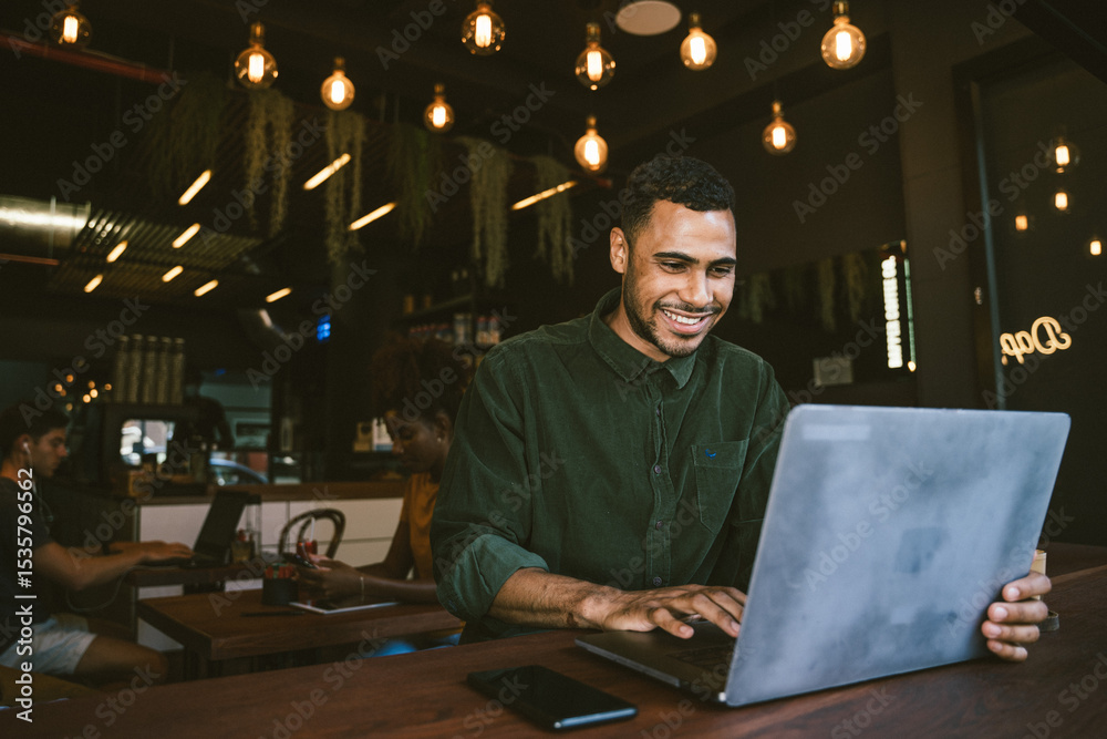 Fototapeta premium Smiling young man typing on laptop while working remotely in cafe