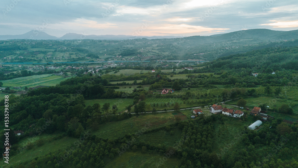 Fototapeta premium Aerial view of green rural landscape with red-roof houses surrounded by fields and orchards in Serbia at sunset