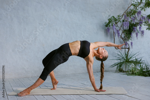 Fotografie Athletic woman in black sportswear practicing a wild thing yoga pose on a mat, arching back gracefully, demonstrating strength, flexibility, and focus in a serene, well-lit indoor space