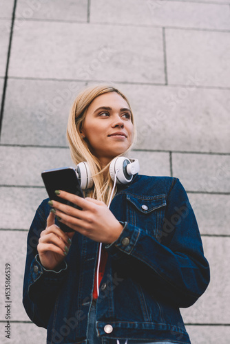 Below view of attractive female blogger with modern headphones publishing new music post on website on digital smartphone via 4G internet connection standing ourdoors on grey promotional background photo
