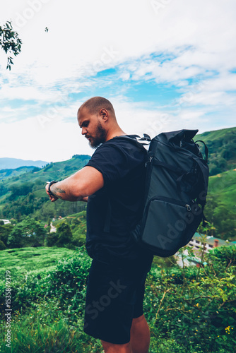Pensive bearded tourist with backpack looking at smartwatch to tracking GPS walking in green locality of asia mountains.Hiker with rucksack managing tame on wristwatch during adventure trek