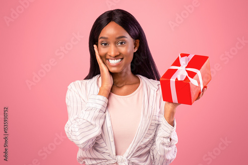 Portrait of suprised African American woman with wrapped gift box posing on pink studio background. Poitive black young lady celebrating special holiday, receiving unexpected present, feeling happy